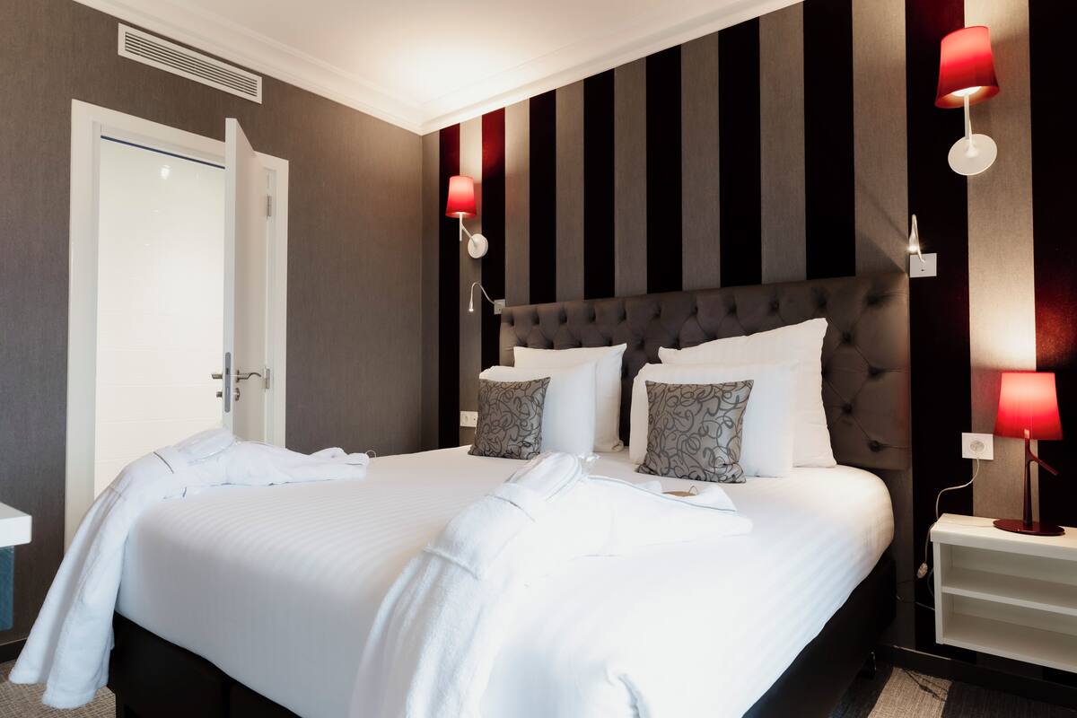 Een net opgemaakt bed met witte lakens en twee gestreepte kussens, een donker bekleed hoofdbord en twee rode wandlampen.