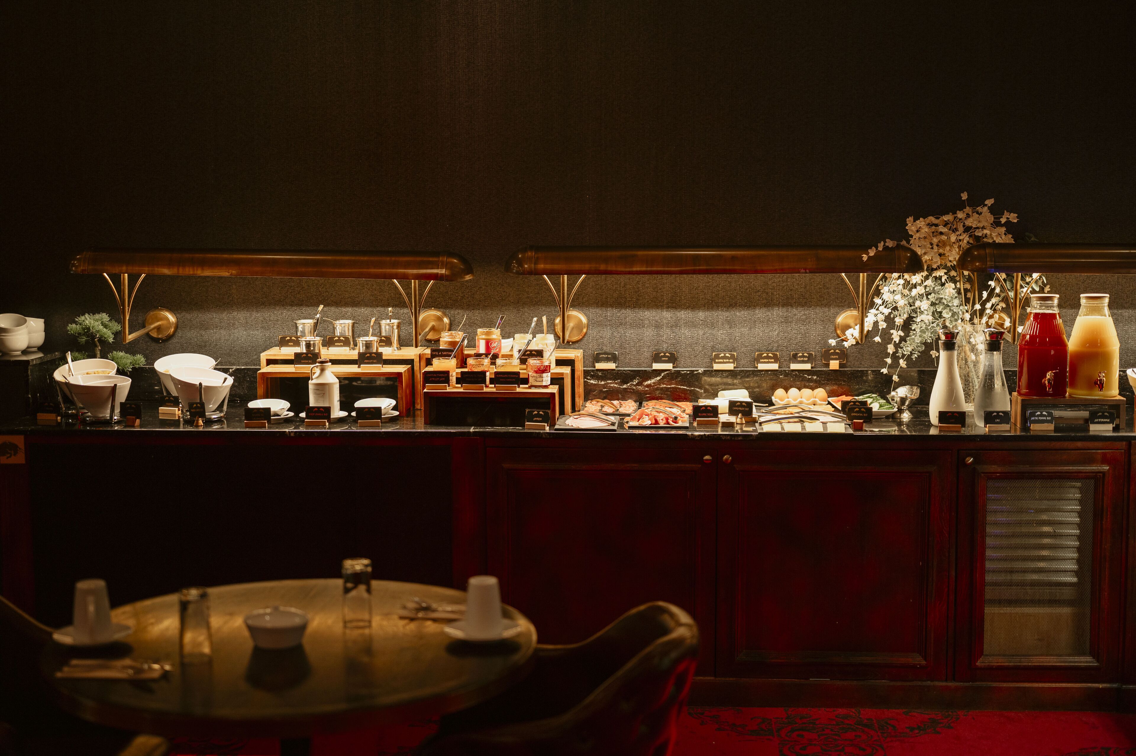 Un espace bar avec un comptoir où sont exposées diverses boissons et denrées alimentaires, une petite table avec des chaises et un vase décoratif rempli de fleurs.