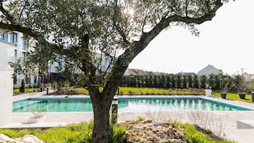 Una piscina con un gran árbol en primer plano, rodeada de vegetación y un edificio moderno al fondo.