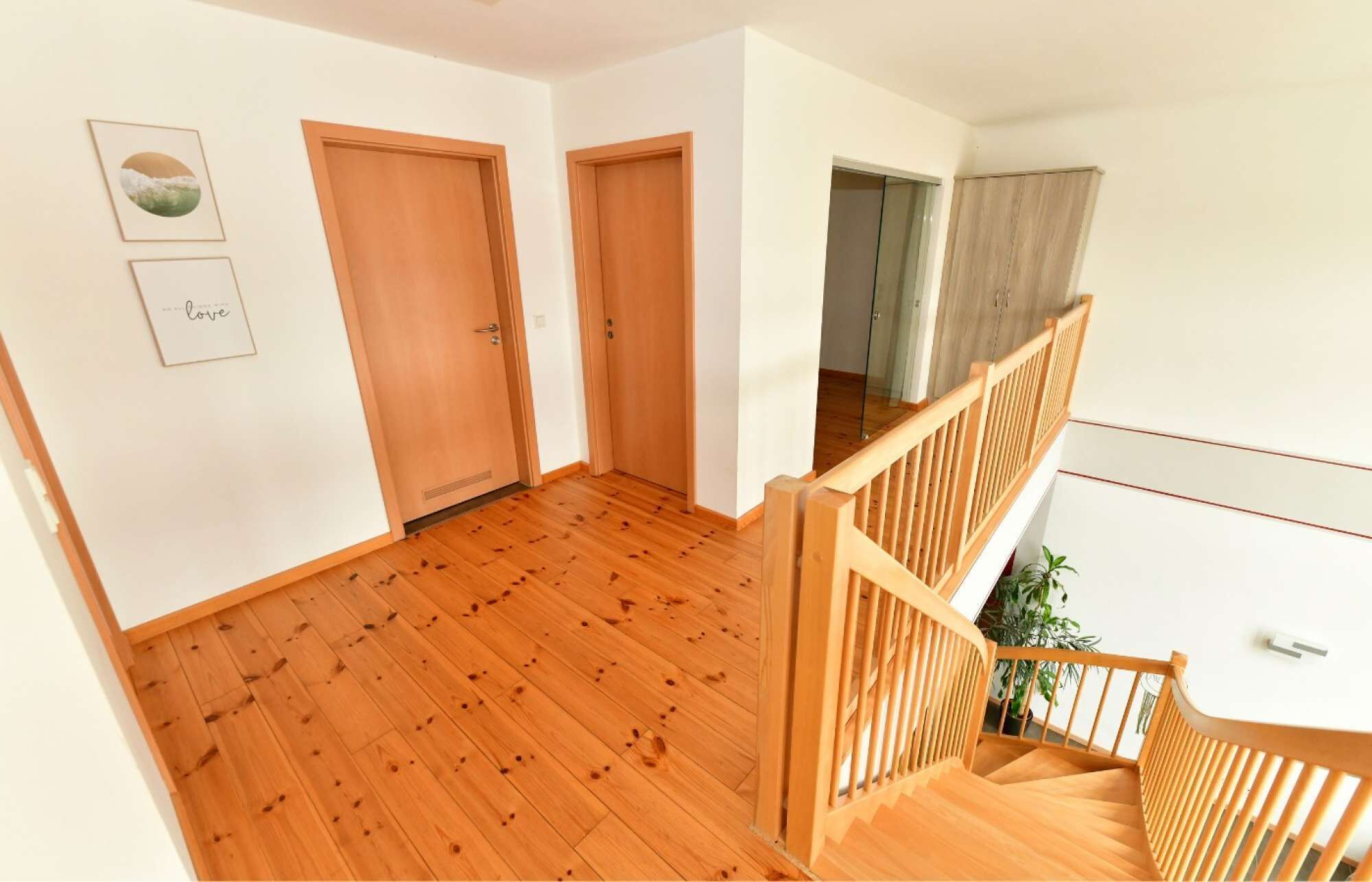 A wooden-floored hallway with two doors, a staircase with wooden railings, and a plant on the landing.