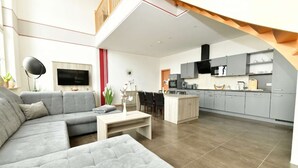 Een moderne woonkamer met een grijze modulaire bank, een salontafel en een televisie. De keuken heeft grijze kastjes, een magnetron en een fornuis.