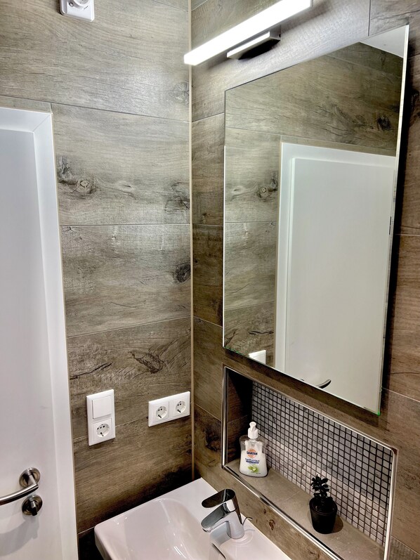A bathroom with a wooden wall, a white sink, a mirror, and a hand soap dispenser.