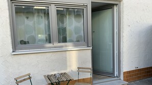 Una petita zona d'estar a l'exterior amb una taula i dues cadires, una finestra amb vidre mat i una porta amb persianes.
