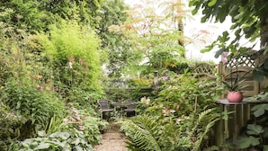 Ein Gartenweg führt zu einer Sitzgruppe mit Tisch und Stühlen, umgeben von üppiger Bepflanzung und verschiedenen Pflanzen.
