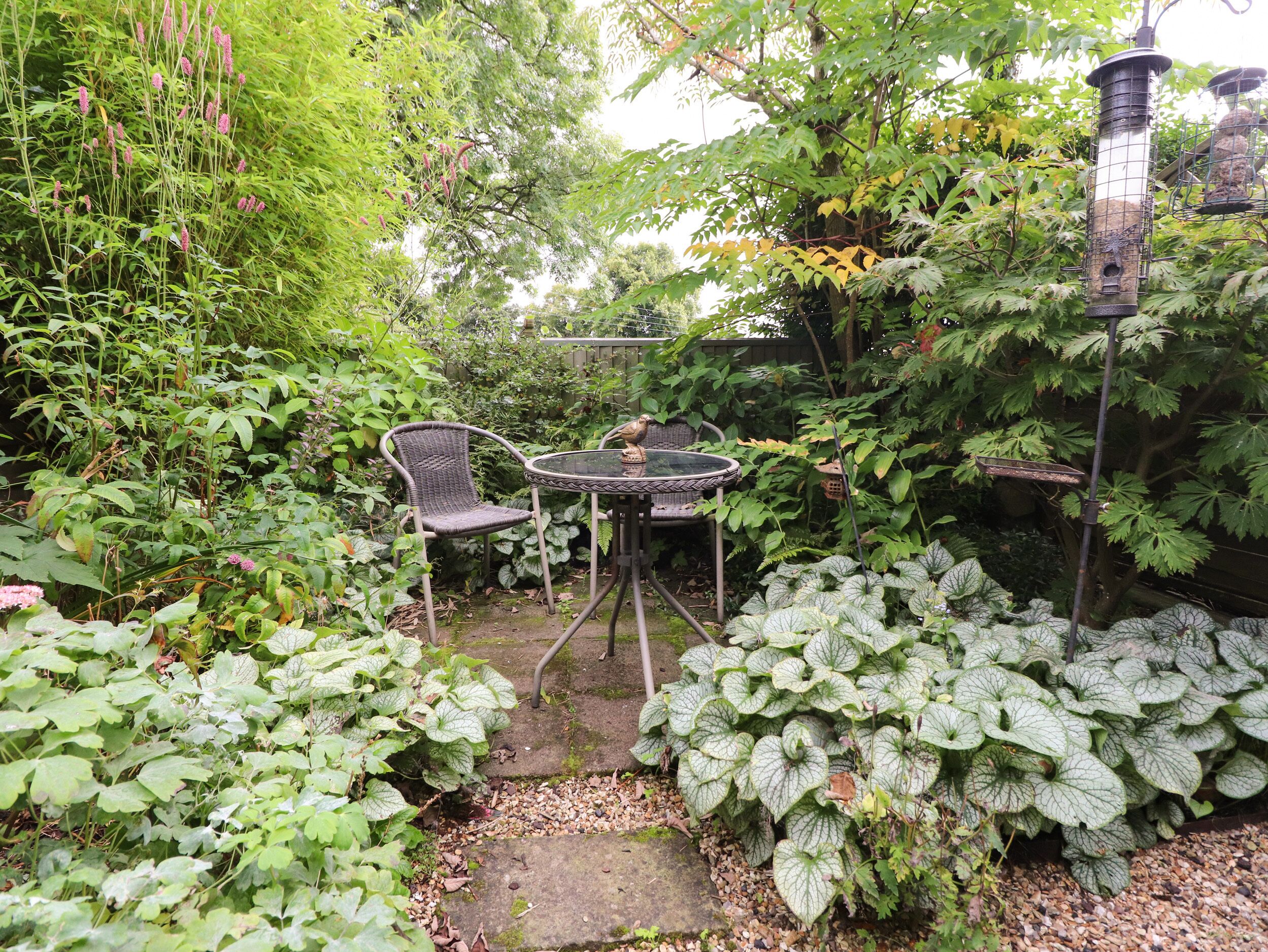 A garden patio with a table and chairs, surrounded by lush greenery and a bird feeder.