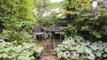 A garden patio with a table and chairs, surrounded by lush greenery and a bird feeder.