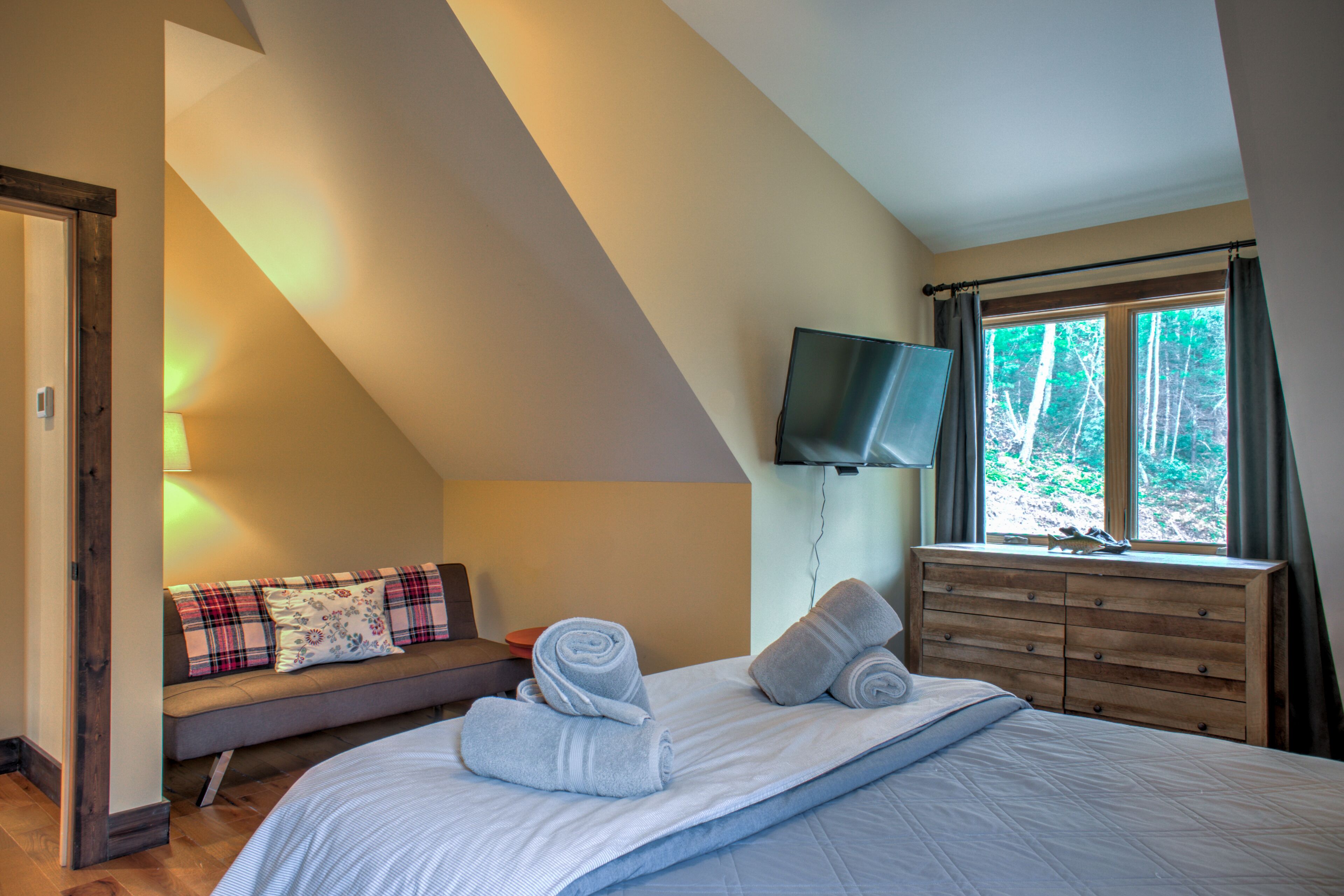 Un dormitorio con cama, cómoda de madera, televisor montado en la pared y ventana con cortinas.
