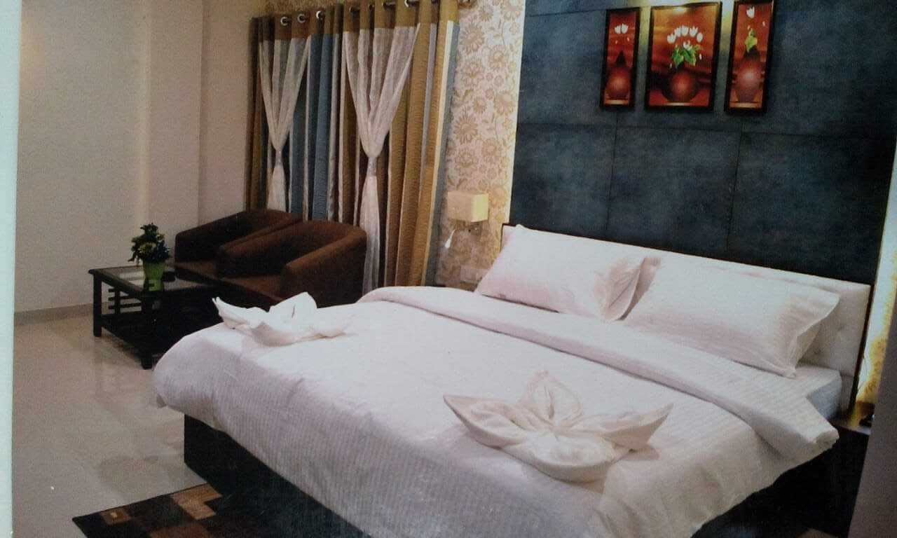 Een netjes opgemaakt bed met witte lakens en decoratieve kussens, een bruine fauteuil, een klein tafeltje met een plant en een wand met drie ingelijste foto's.