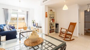 A modern living room with a glass coffee table, a blue sofa, a wooden rocking chair, and a bookshelf. - Duplex Algarrobo Costa Solset (Algarrobo)