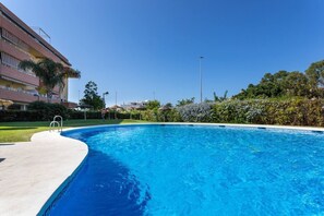 Une piscine d’un bleu éclatant, bordée d’une terrasse en béton, entourée de verdure et d’un bâtiment doté de balcons.
