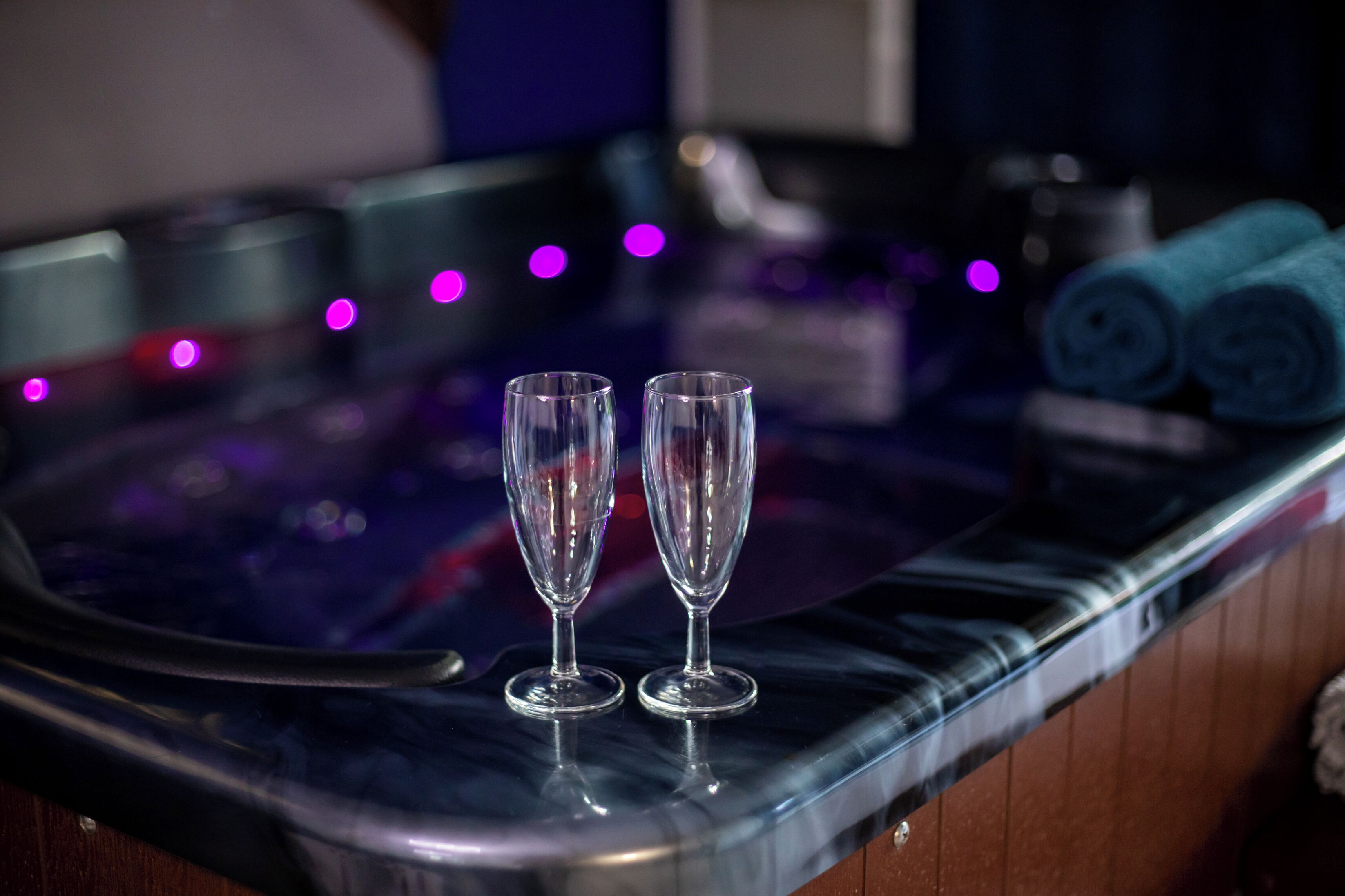 A spa area with two empty glasses on a reflective surface, a towel rack with rolled towels, and a small table with a bottle and a cup.