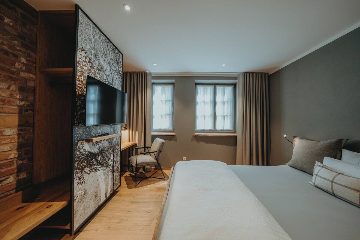A modern hotel room with a large bed, a desk with a chair, a TV mounted on a wall, and two windows with curtains.