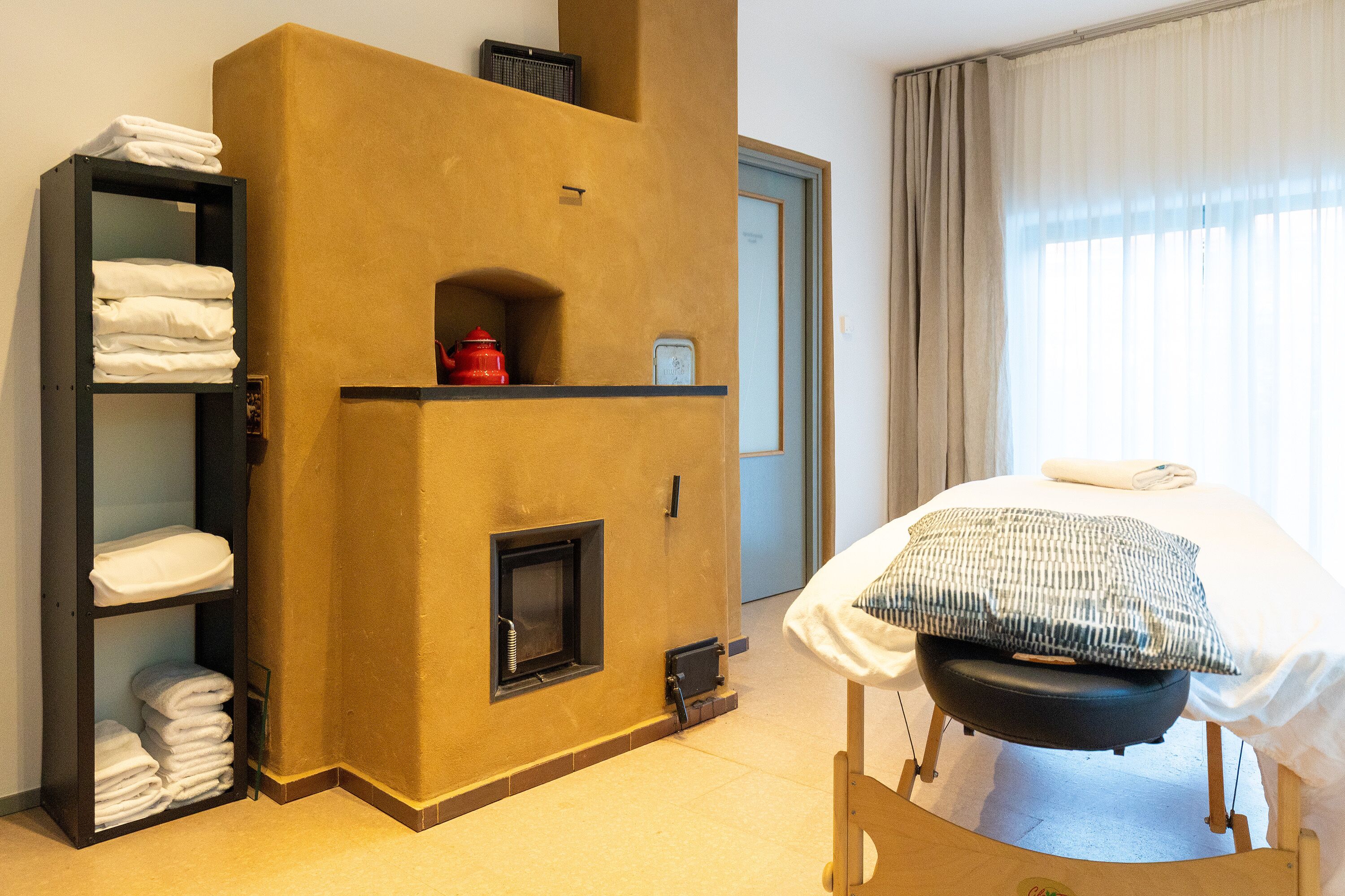 A hotel room with a built-in fireplace, a bed with a pillow, a shelf with folded towels, and a door.