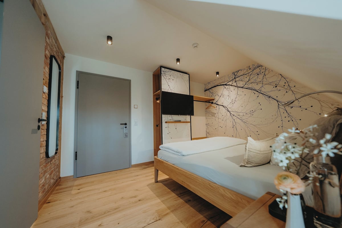 A modern bedroom with a wooden floor, a bed with white bedding, a TV mounted on the wall, and a decorative wall piece.
