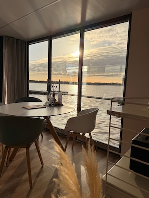 Een moderne kamer met een groot raam met uitzicht op een waterpartij, een ronde tafel met stoelen en een zicht op verre gebouwen.