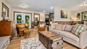 A spacious living room with a sofa, armchairs, and a wooden coffee table. There is a fireplace, a dining area, and a kitchen visible in the background. - Mt. Index Riverside with hot tub, wifi, grill, fireplace and more! (Gold Bar)