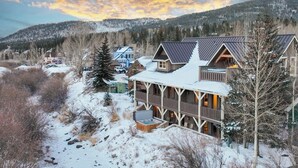 Un gran albergue con un porche cubierto y numerosas ventanas, rodeado de nieve y árboles, con montañas al fondo.
