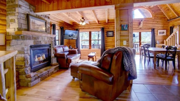 Interior acogedor de una cabaña con chimenea de piedra, sofá de cuero, mesa de comedor de madera y una escalera que conduce a un nivel superior.