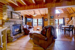 L’intérieur chaleureux d’un chalet avec une cheminée en pierre, un canapé en cuir, une table à manger en bois et un escalier menant à un étage supérieur.
