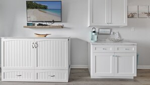 Ein Zimmer mit einem großen Flachbildfernseher, der eine Strandansicht zeigt, weißen Schränken mit Schubladen, einer Marmorarbeitsplatte und einer Kaffeemaschine.