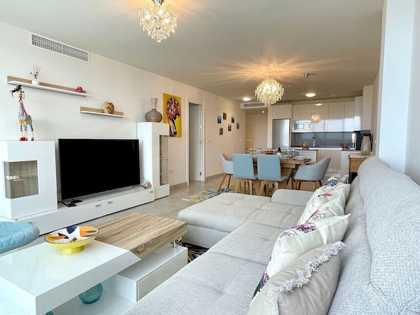 A modern living room with a flat-screen TV, a sofa, a dining area, and a kitchen in the background.