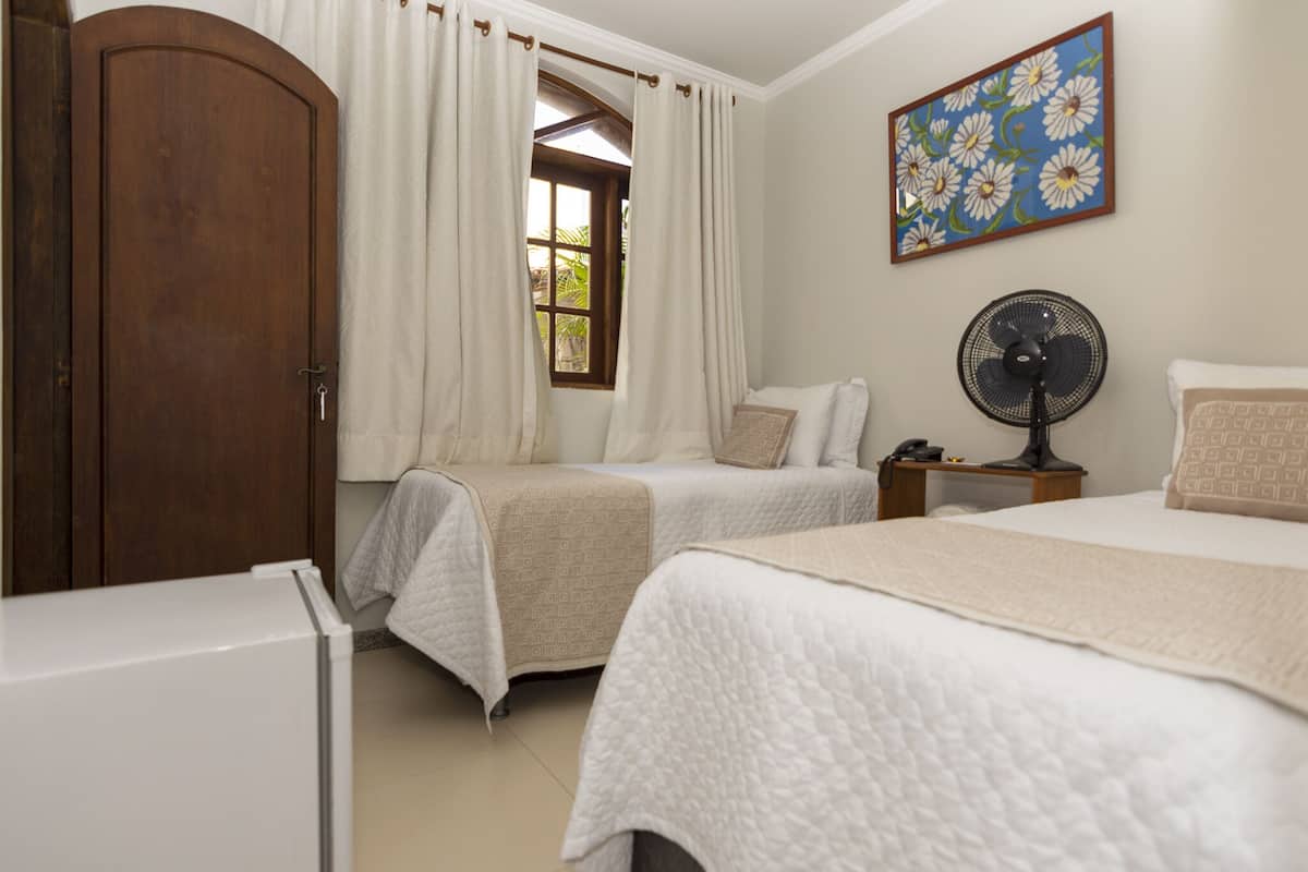 A bedroom with two beds, a wooden wardrobe, a window with curtains, a fan, and a painting on the wall.