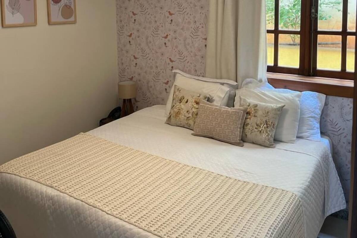 A bedroom with a large bed, a hanging light fixture, a window with curtains, and two framed pictures on the wall.