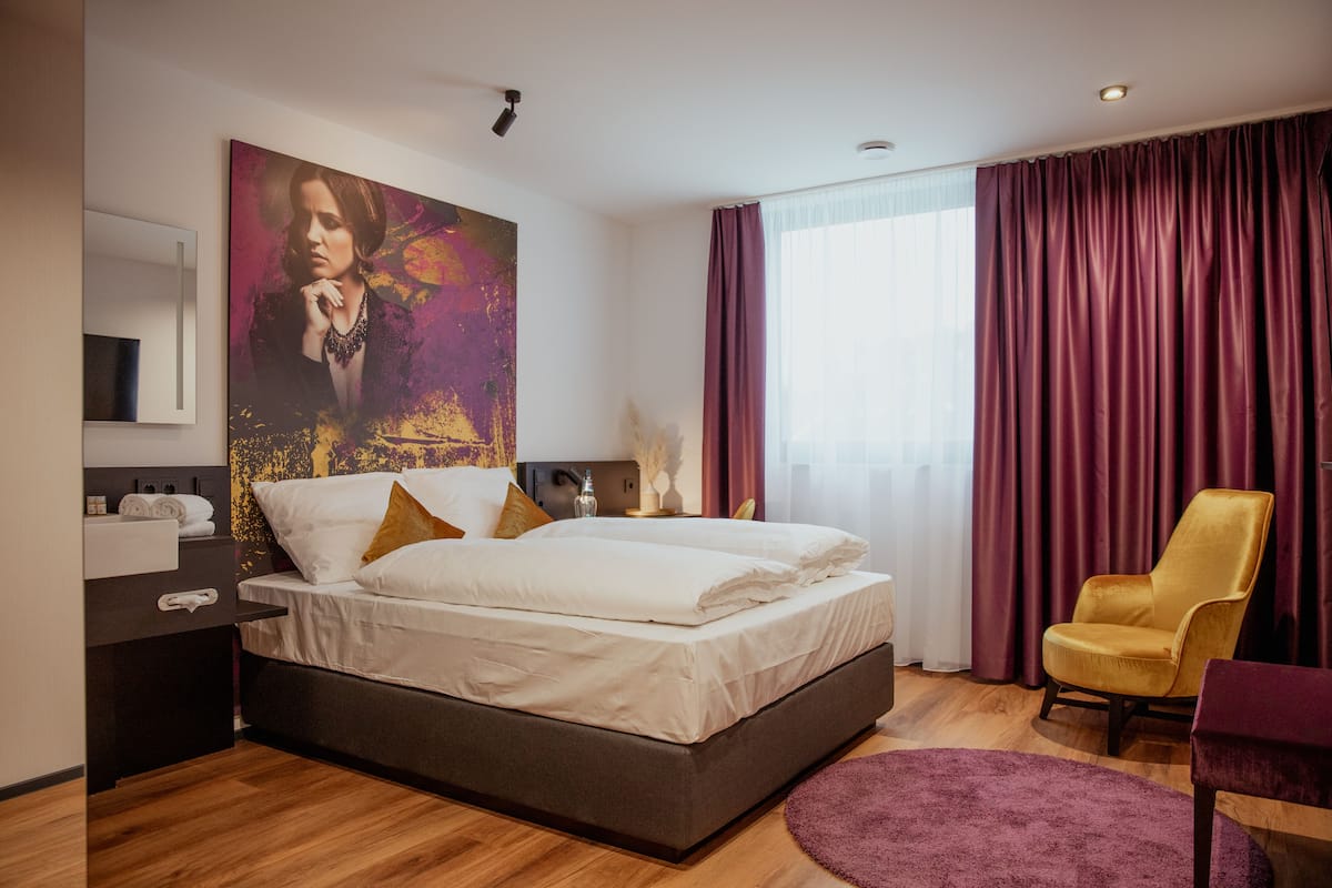 Ein modernes Hotelzimmer mit einem großen Bett, einem Schreibtisch mit Stuhl, einem großen Fenster mit Vorhängen und einem Gemälde an der Wand.