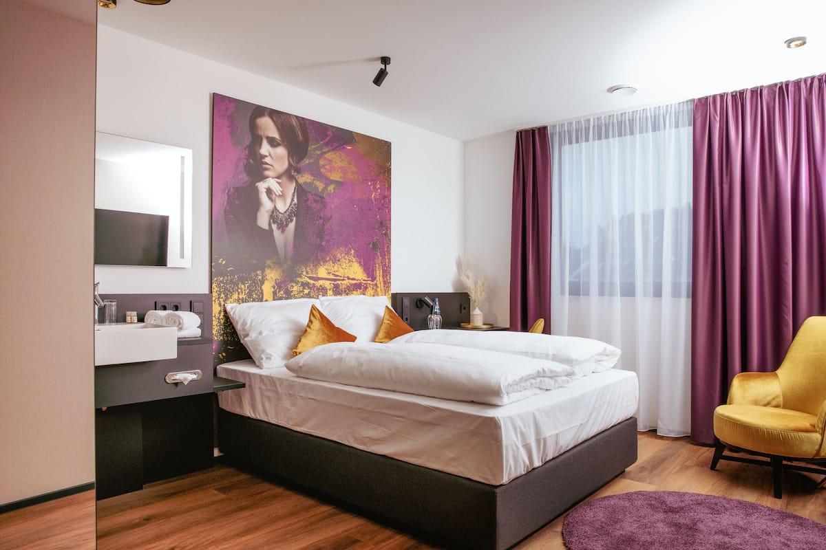 Ein modernes Hotelzimmer mit einem großen Bett, einem Nachttisch mit Lampe, einem Badezimmer mit Waschbecken und Spiegel sowie Blick auf die Stadt durch große Fenster mit violetten Vorhängen.