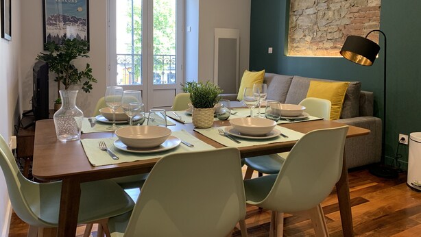 A dining area with a wooden table set for four, surrounded by chairs. A large window with a view of greenery is visible, and a stone wall with a lamp is in the background. - #CD L' Ainé 👨‍👩‍👦‍👦| Jean Jaurès | Tramway A, B and E| T3 équipé (Grenoble)