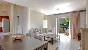Una sala de estar moderna con una mesa de comedor, sofá y mesa de centro. Hay una zona de cocina con armarios y un refrigerador. Un ventanal proporciona luz natural y se ve una planta al fondo.