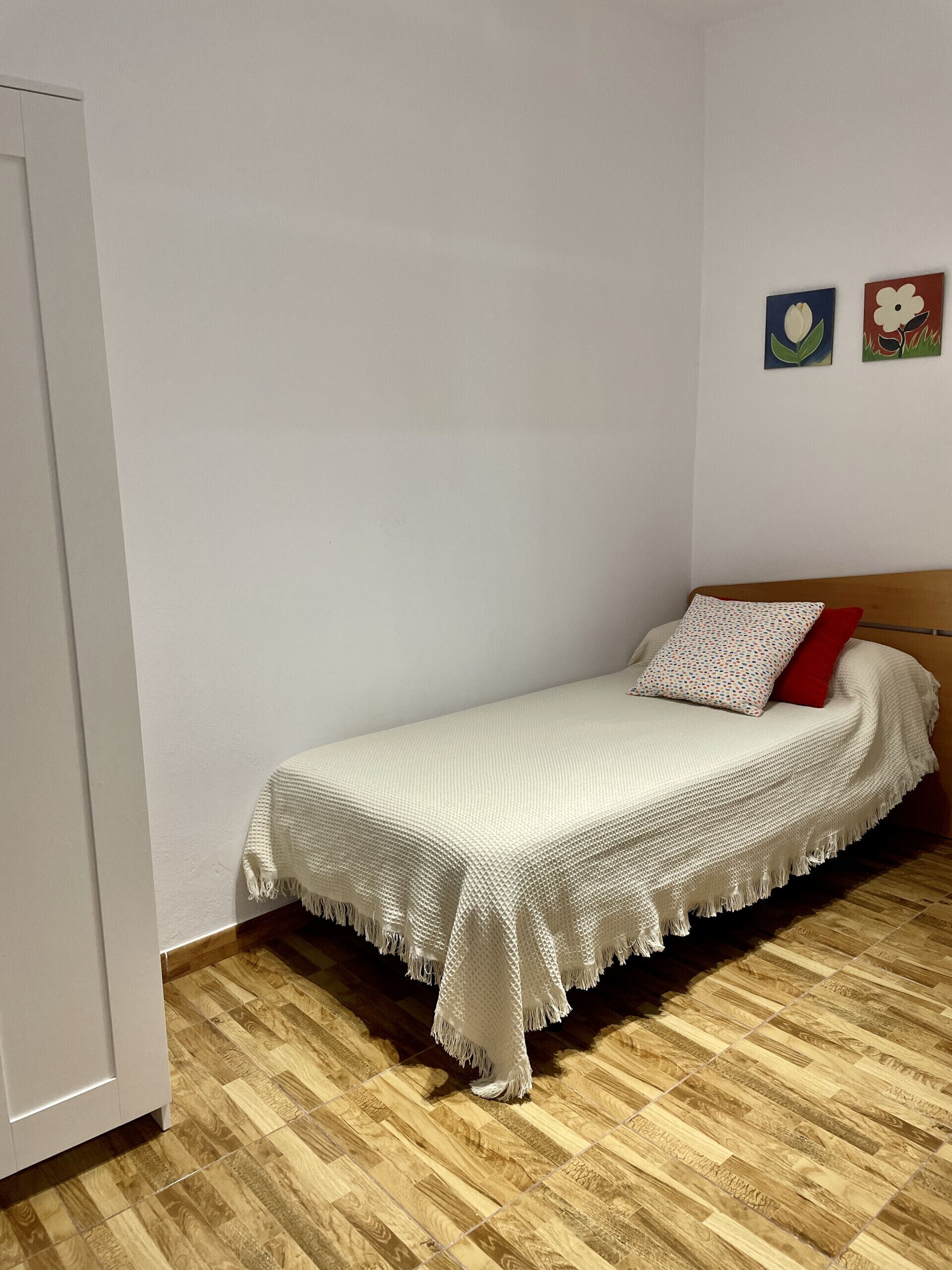 A single bed with a white blanket and two pillows, a wooden floor, and a white wall with two small artworks.