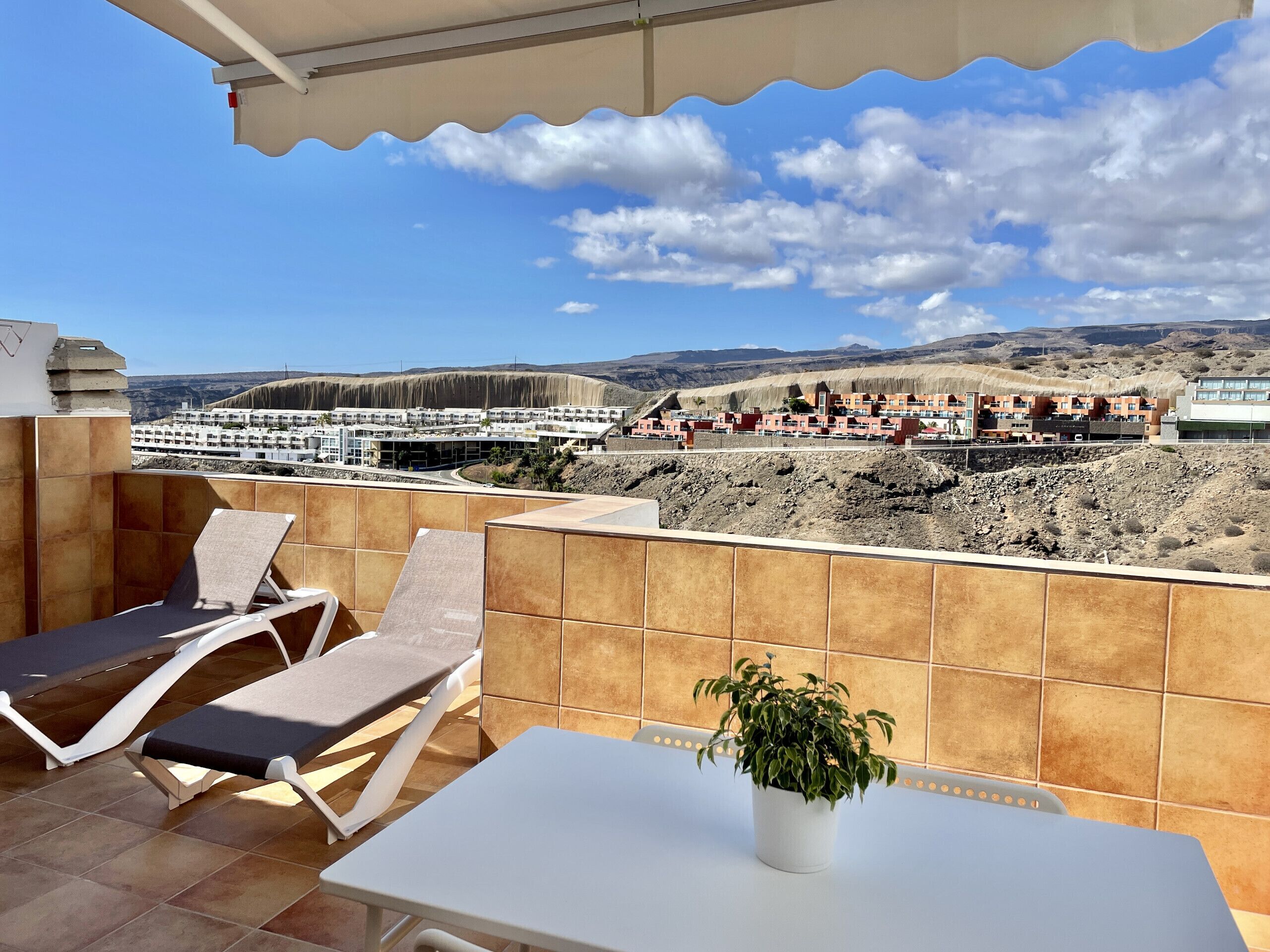 A terrace with two lounge chairs and a table, a potted plant, and a view of a coastal town with buildings and hills.