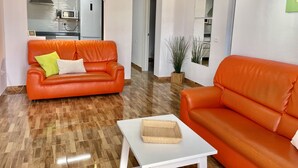 Uma sala de estar moderna com dois sofás laranja, uma mesa de centro branca e uma cozinha visível por um arco.