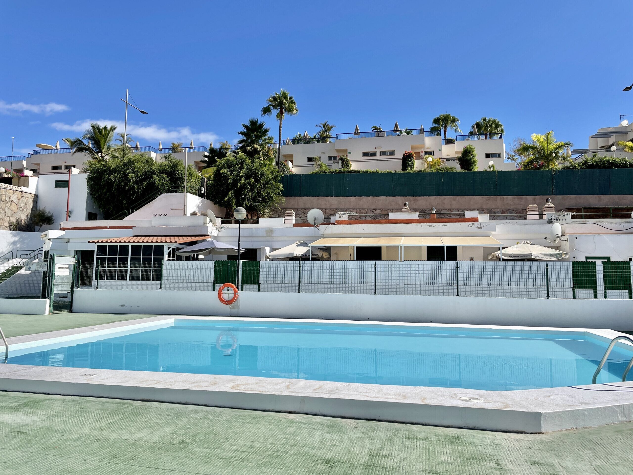 A swimming pool with a clear blue water surface, surrounded by a white pool deck. In the background, there are buildings with balconies and satellite dishes, and a green fence separating the pool area from the buildings.