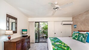 A bedroom with a bed, a TV, a ceiling fan, a dresser, and a view of a garden through a glass door.