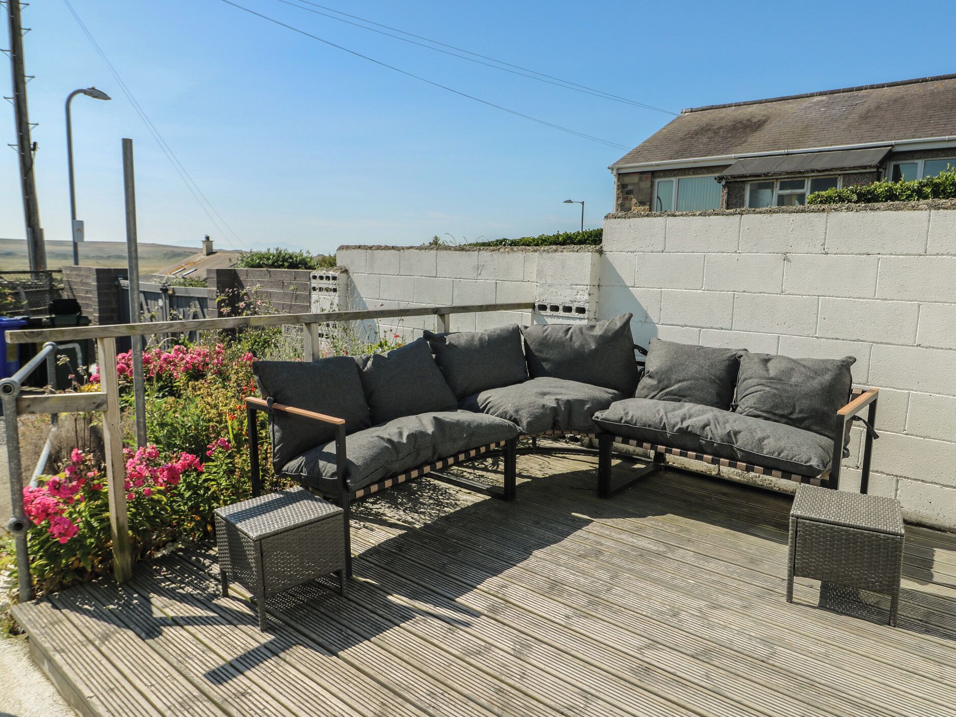 A patio with a grey sofa, two armchairs, and a small table, surrounded by a wooden deck and a concrete wall.