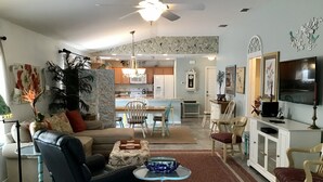 Uma sala de estar espaçosa com um sofá de couro preto, uma área de refeições com mesa de madeira e uma cozinha ao fundo.