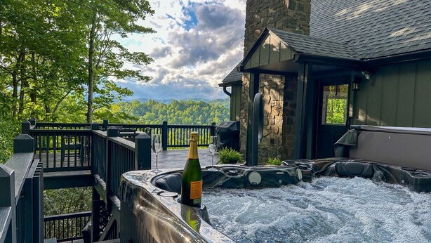 Un espace avec un jacuzzi, une bouteille de champagne, une terrasse en bois et un bâtiment en pierre offrant une vue sur la forêt.