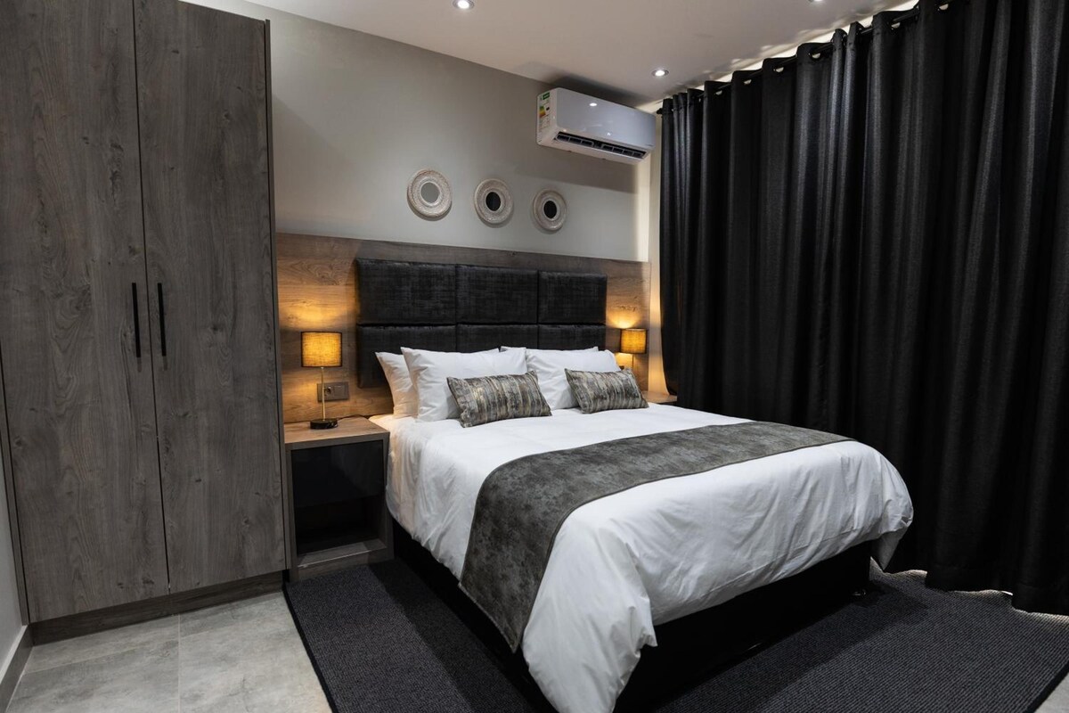 A modern bedroom with a bed, bedside lamps, an air conditioning unit, and a dark curtain.