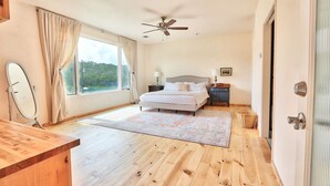 Une chambre à coucher avec un lit, un ventilateur de plafond, une fenêtre avec des rideaux, une table de chevet avec une lampe et un plancher en bois.