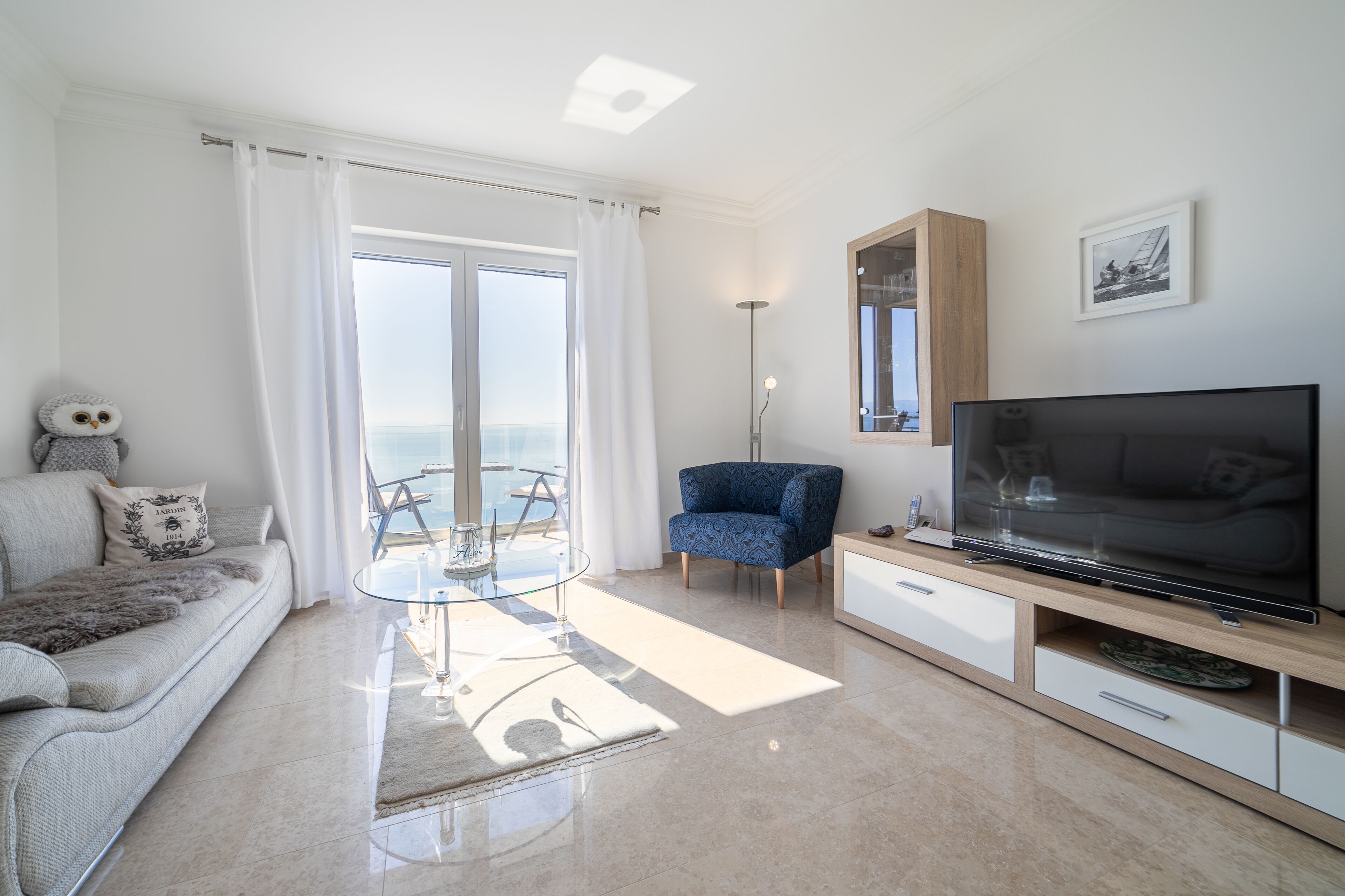 Un salon moderne avec un canapé, une table en verre, un fauteuil bleu, un téléviseur et une vue sur la mer.