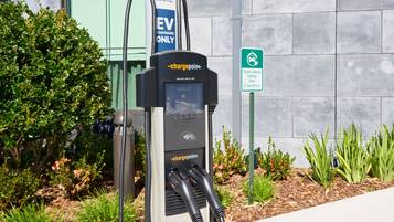 An EV charging station with a digital display, located outdoors with a building and greenery in the background.