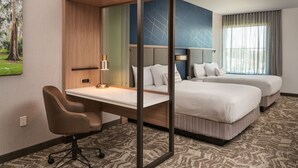 A hotel room with a desk, two beds, a chair, a lamp, and a painting on the wall.