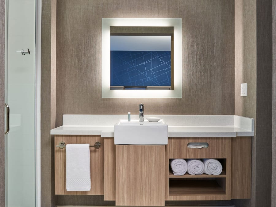 A modern bathroom with a glass shower door, a white sink, a mirror with a geometric wall art, and wooden cabinets.