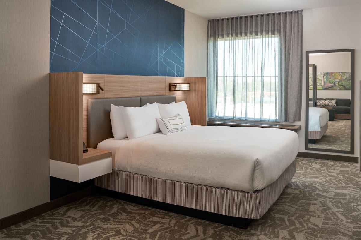 A hotel room with a large bed, a nightstand, a wall-mounted lamp, and a large window with curtains.