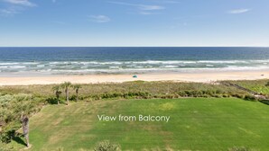 Une vue depuis un balcon donnant sur une plage, avec des vagues, une étendue de sable et quelques zones verdoyantes.