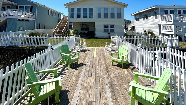 A wooden deck with green Adirondack chairs, a white picket fence, and a two-story building with a balcony in the background. - Canal home, 3 bedrooms, 2 bathrooms, dock, pet friendly, fenced in back yard. (Supply)