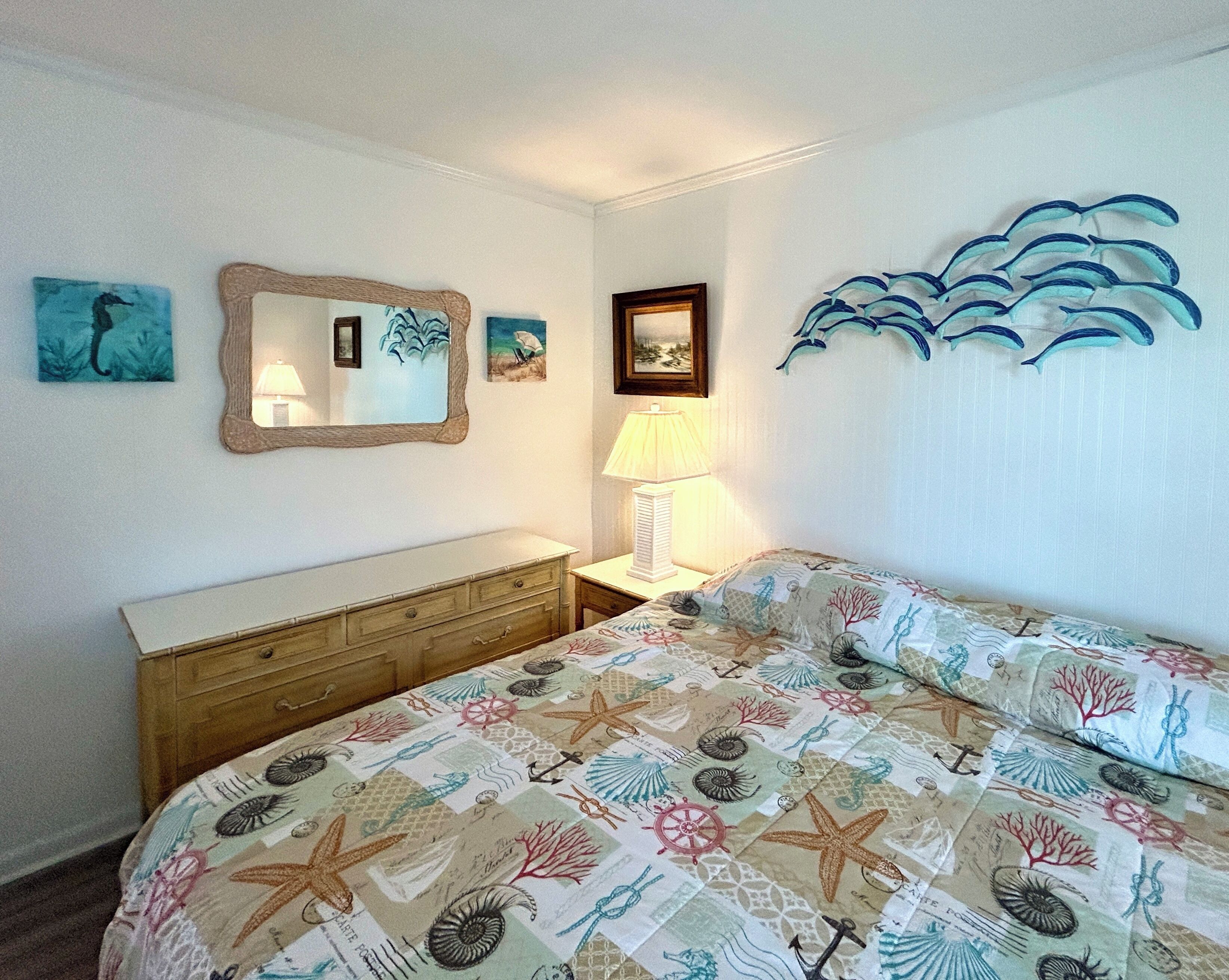 A bedroom with a bed, a dresser, a nightstand, a lamp, and wall art including a mirror, framed pictures, and a seascape mural.