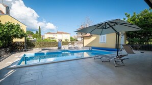 Uma piscina com espreguiçadeiras e um guarda-sol, cercada por um terraço ladrilhado e um sebedeiro.